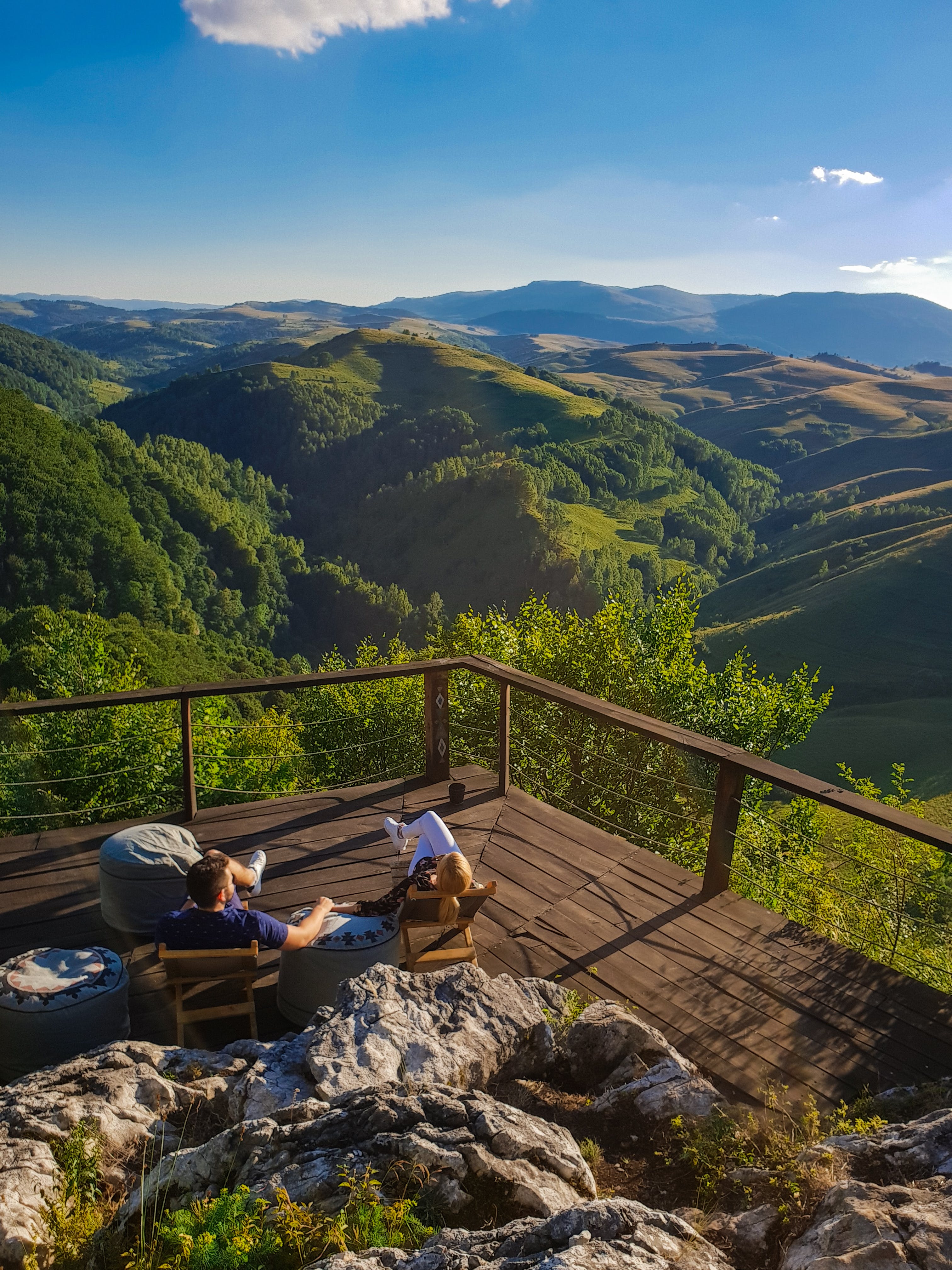 High Angle Photo Of A Man And Woman Sitting on Balcony Viewing Mountains Under Blue and White Sky